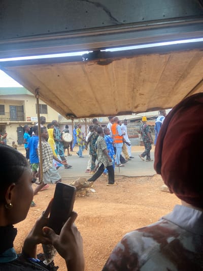 In Okengwe - Obehira, Nigeria, a procession of people walks on a paved road, some holding or playing drums, seen from an obscured viewpoint through a building structure. Some are wearing traditional or brightly colored clothing. An individual holds a mobile phone, partially obscuring the view of the procession. Buildings can be seen in the background.