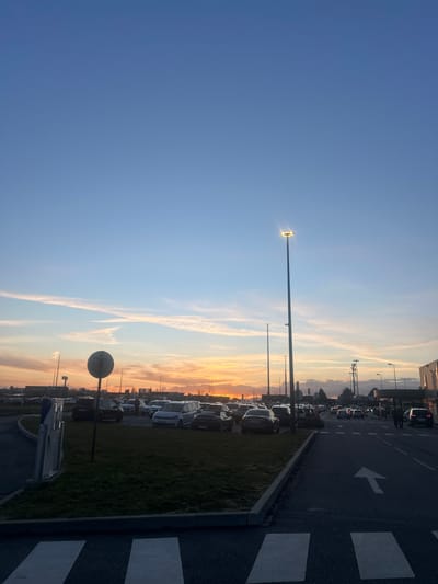 The image shows a parking lot at dusk, with cars lined up next to a grassy median and an adjacent road. A directional arrow is painted on the road's surface. A tall street lamp illuminates the scene. The sky transitions from blue to a mix of orange and yellow hues as the sun sets, creating a striking backdrop with some thin clouds. The image appears to be taken from a low angle.