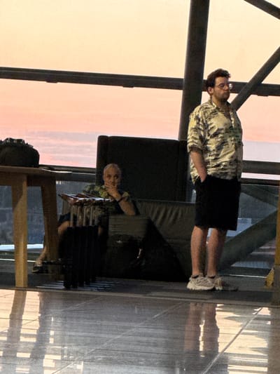 The image captures a scene inside what appears to be an airport terminal in Zaventem, Belgium. In the foreground, a person stands with their hands in their pockets, wearing a patterned short-sleeved shirt and dark shorts. They are positioned near a window that offers a view of a soft, pinkish sky, possibly indicating sunset or sunrise. To the left, another person is seated, partially obscured by a dark chair, and appears to be resting their chin on their hand. Luggage is visible near the seated individual. The environment features modern architecture with prominent structural beams and a reflective floor. The lighting suggests an indoor setting with natural light filtering in from the large windows.