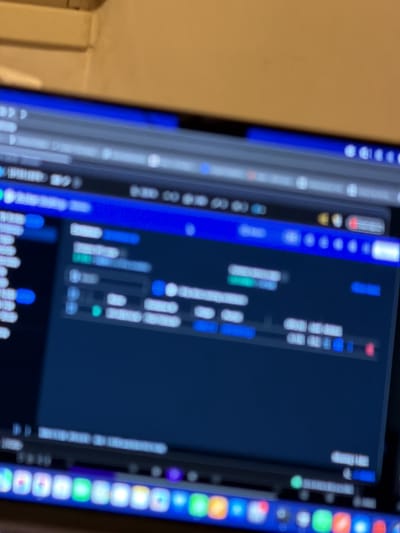 The image depicts a close-up, out-of-focus view of a computer monitor displaying a graphical user interface, observed indoors in Kafr Yasif, Israel. The monitor screen is illuminated, showing a predominantly dark-themed interface with blue and grey elements, along with scattered lighter areas representing unreadable text, icons, or interface components. A row of colorful application icons, consistent with a taskbar or dock, is visible along the bottom edge of the screen. The entire display is blurred, precluding the identification of specific software, content, or textual information. A light-colored wall or surface is visible in the blurred background above the monitor. No individuals are present in the frame, and no specific actions or interactions are discernible beyond the computer being powered on and displaying a user interface.