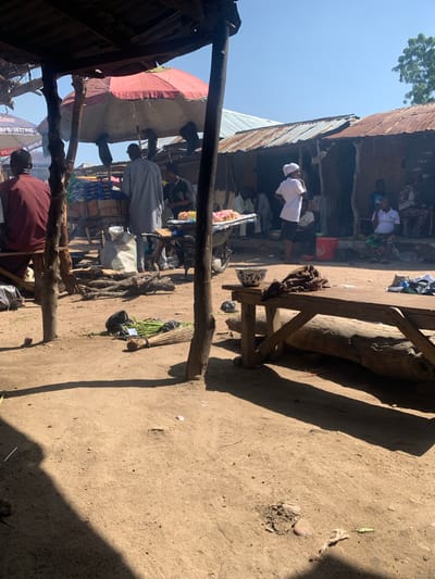 The image depicts a bustling market scene in Boi A, Nigeria. The scene is outdoors, with a dirt ground and simple, rustic structures that suggest a traditional marketplace. The overhead lighting and long shadows indicate it is daytime, likely late morning or early afternoon. The weather appears to be clear and sunny.

Several individuals are visible, dressed in everyday clothing, some wearing white garments and head coverings, suggesting participation in a religious or cultural observance. Some people appear to be vendors, while others are customers or passersby. The activity suggests a typical market day, with goods being displayed and transactions likely taking place.

Objects in the scene include various stalls with goods, a large red container, a broom made of natural materials, and wooden structures that serve as part of the market's infrastructure. One stall is covered by a large, red umbrella. A large tree trunk has been incorporated into a wooden bench or table.

Notable details include the corrugated metal roofing of some structures, indicative of makeshift or semi-permanent market stalls. The overall atmosphere seems lively and busy, typical of a community gathering point. There are no overt signs of distress or negative emotions; the scene appears to be one of everyday commerce and social interaction.

There is some visible text, though it is difficult to read clearly. One sign, partially obscured, appears to have English words such as "SETTING" and "RAPID," which could relate to business names or product advertisements.