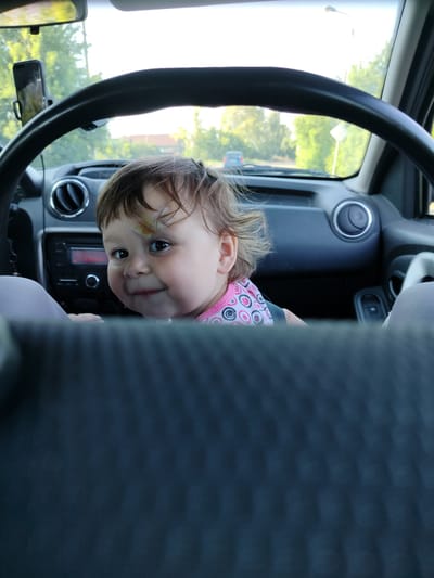 A close-up shot captures a toddler sitting in the driver's seat of a car, as if steering. The child, with a bandage on their forehead, looks directly at the camera with a curious expression. The toddler wears a pink shirt with a circular pattern. The car's dashboard is visible, including the steering wheel, air vents, and radio. Through the windshield, a blurred outdoor scene shows trees and buildings, suggesting daytime. The lighting and the children's attire imply it's a warm day. The image appears to be taken from the back seat of the car.
