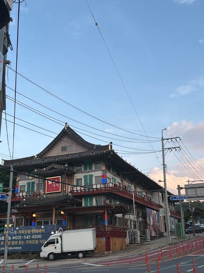The image depicts a multi-story building with traditional Korean architecture in Pocheon-si, South Korea. The building features dark tiled roofs with upturned eaves and wooden detailing. It appears to be a restaurant or similar establishment, as indicated by signage and decorative elements. A white box truck is parked in front of the building. The scene is set on a street with power lines crisscrossing overhead. The sky is partly cloudy with the light suggesting it is either early morning or late afternoon. The street has painted lanes and traffic cones, indicating an urban environment.