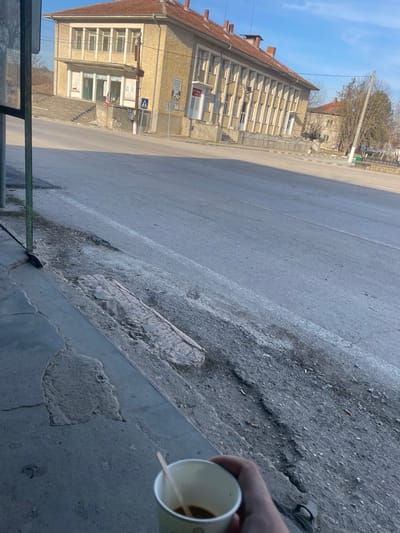 This image depicts a daylight street scene in Topchii, Bulgaria, under a clear blue sky. In the immediate foreground, a right human hand with light skin tone holds a white disposable cup containing a dark liquid and a wooden stir stick. The hand is positioned over a gray concrete sidewalk exhibiting cracks and patches of repair. To the left, a dark green metal pole stands vertically. The edge of the sidewalk transitions to a rough, uneven dirt and gravel shoulder beside an asphalt road. This shoulder contains exposed soil, scattered debris, and several discarded cigarette butts.
Across the asphalt road, a two-story building with a light yellow/beige facade and a red tiled roof dominates the midground. The building features multiple rectangular windows on both levels. The ground floor has several entrance areas, one with a set of concrete steps leading up to it, and another section with prominent vertical concrete elements. Road signs, including a pedestrian crossing sign and directional markers, are visible on poles along the street. Further down the street to the right, a second, similar building is partially visible, accompanied by several bare trees, suggesting a non-summer season. The street and surrounding area are devoid of human activity or vehicles.