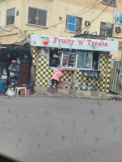 This media file captures a street scene in Alagbede, Nigeria, likely during daytime given the lighting. The focal point is a small shop or food stall named "Fruity 'n' Treats," distinguished by a prominent sign with a fruit logo and red lettering. The shop's facade is decorated with a checkered pattern of yellow and black tiles.

In front of the shop, two individuals are visible. On the left, a person is standing near a collection of items, including sacks and buckets, appearing to be engaged in some activity. To the right, another person, wearing a pink top and dark leggings, is also occupied with an activity, possibly cleaning or preparing something. Behind them, a display case shows various food items, suggesting the stall serves shawarma, snacks, parfaits, and juice, as indicated by the text on the sign.

The building behind the shop has multiple windows, some with security bars. Electrical wires crisscross above the scene, a common sight in many urban environments. The street in the foreground is wet and reflective, hinting at recent rain or possibly just a damp surface. The overall atmosphere appears to be that of a bustling local business operating in a busy street. Visible text on the sign includes the business name, phone numbers (09056203098, 08132730434), and an email address (Freyandtreats@gmail.com). A small sign for "PHOTO STUDIO P" is also visible on the building's upper level.