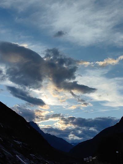 The image depicts a mountainous landscape in Damphug, India, under a dramatic sky filled with clouds. The foreground is dominated by the dark silhouettes of two steep mountain slopes, framing a valley. The left slope is darker and more textured, appearing closer, while the right slope is slightly less dark and reveals layered mountain peaks in the distance.

The sky is a canvas of blues and grays, with scattered clouds of varying density. Some clouds are dark and heavy, suggesting impending weather, while others are lighter, allowing patches of sunlight to break through, illuminating the edges of the clouds with a warm, golden hue. The interplay of light and shadow across the sky and mountains creates a sense of depth and atmosphere.

The environment appears natural and rugged. There is no visible human presence or activity in the immediate scene, nor any prominent man-made structures other than faint, distant indications of what might be lights or structures at the very bottom right, and possibly some infrastructure elements at the bottom center of the image, obscured by the darkness. The focus is on the grandeur of the natural scenery and the dynamic sky. The overall impression is one of a remote, awe-inspiring mountain vista.