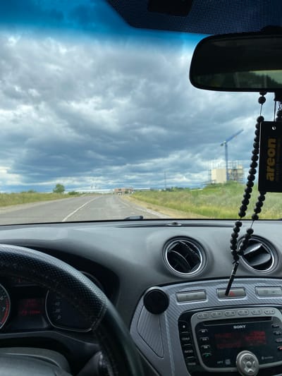 The image, taken from the interior of a car, shows a cloudy sky with dark, heavy clouds. In the distance, a construction site is visible with a blue crane and incomplete structures. The road ahead appears to be asphalt and is slightly winding. On the dashboard, a car's steering wheel is visible on the left, with the instrument cluster showing "195 km". To the right of the steering wheel, the car's center console has an air conditioning vent and a Sony car stereo. A car air freshener with black beads and the "areon" logo hangs from the rearview mirror. The scene suggests a drive on the outskirts of Grătiești, Moldova, possibly on a cloudy day. There are no people visible in the image.