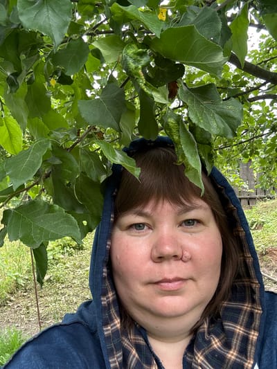 A close-up, waist-up selfie of a woman is captured outdoors, with lush green leaves of a tree partially obscuring the top of the frame. The woman, with medium-brown hair, wears a dark blue hooded sweatshirt with a plaid lining visible around the hood and collar. Her head is slightly tilted, and she is looking directly at the camera. She has fair skin, noticeable redness on her cheeks, and grey-blue eyes. A raised, flesh-colored growth is visible on the bridge of her nose. The background consists of a grassy yard with a weathered wooden fence visible in the distance. The overall setting suggests a rural or suburban environment, possibly a backyard in Ильпанур, Russia, given the presence of deciduous trees and a rustic fence.