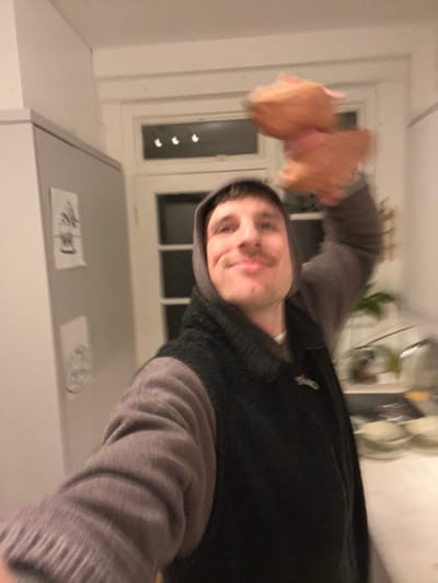 A man poses in a kitchen, holding a blurry pink stuffed animal in his right hand. He wears a grey hoodie over a black vest. White cabinets are above him with papers taped on. A white window frame is visible behind him, providing light. The photo is blurry and slightly tilted. Kitchen items can be seen on a white countertop below.
