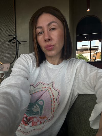 A woman is depicted in a close-up selfie, centrally framed, seated indoors in what appears to be a cafe or restaurant in Усады, Russia. She has medium-brown, bob-cut hair with subtle lighter streaks visible, particularly on the left side of the frame. Her eyes are open, with dark irises. Her lips are pursed in a pouting expression. She is wearing a white long-sleeved top featuring a multi-colored graphic design on the chest, which includes abstract shapes and the partial text "il Cielo Stellato." The fabric of the top has a textured, slightly sheer appearance.
The indoor background features walls with a dark greenish-grey, textured finish. Two arched openings are visible: one to the left and one to the right of the woman. To the left, behind her, a dark metal grid-like panel and a black garment rack are visible. Further left and slightly behind, a light pink baby high chair is partially visible. To the right, through the archway, a multi-paned window provides a view of an outdoor scene. A red cylindrical light fixture emitting a warm glow hangs from the ceiling above the right arch. The woman is seated on dark green upholstered furniture, visible in the lower right portion of the frame.
The outdoor view through the window shows a bright sky, green trees, and multiple structures. A light-colored awning or umbrella structure is prominent, along with a building or house-like structure with a dark roof and a white-framed window. A portion of a white vehicle is also visible on the far right of the outdoor scene.