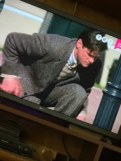 A television screen displays a scene from a movie or show. In the foreground, a man with dark hair is hunched over, wearing a grey tweed suit over a collared shirt and a patterned sweater vest. His right hand, illuminated with a pinkish hue, is extended downwards, possibly touching something on the ground. The background of the screen shows a blurry outdoor setting with a light-colored wall and green foliage. In the upper right corner of the screen, three white circles with blue lettering are visible, forming the logo "TET," a Ukrainian television channel. The LG logo is present in the bottom left corner of the television screen. The television itself is placed on a wooden shelf, with electronic devices and black discs visible on the shelf below. The overall scene is captured from a slightly elevated angle, showing the television screen and its immediate surroundings. The image does not provide information about the specific location within the city of Osolynka, Ukraine.