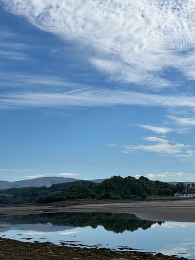 The image depicts a serene natural landscape with a body of water in the foreground reflecting the sky and surrounding trees. In the middle ground, there is a stretch of sandy shore and a dense line of green trees forming a natural barrier. Beyond the trees, rolling hills covered in greenery recede into the distance. A small white building with a dark roof is visible on the right side of the image, near the tree line. The sky is a vibrant blue with scattered white clouds, some appearing wispy and others more textured. The water's surface is calm, creating a clear reflection of the sky and the land. The scene appears to be located in a rural or semi-rural area. The image is not set in the city of Dunuisce, Ireland.
