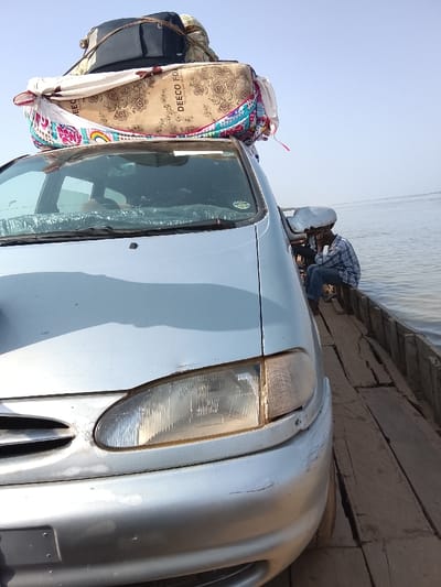 A silver passenger vehicle is depicted loaded onto a wooden ferry, indicating a river crossing in Ibi, Nigeria. The vehicle's roof is heavily laden with luggage, including a dark rectangular bag and a large, bundled item wrapped in patterned fabric, prominently featuring "DEECO FOAM" text and a rainbow design, all secured for transit. The car's front features visible headlights, a silver bumper with minor scuffing, and a hood. A male individual, wearing a blue and white plaid shirt and blue jeans, sits barefoot on the exposed wooden planks of the ferry alongside the car's passenger side, appearing to rest with a hand near his face. The ferry is constructed from rough, parallel wooden planks. A calm body of water extends to the right of the car and behind the individual, meeting a clear, light blue sky under daylight conditions.