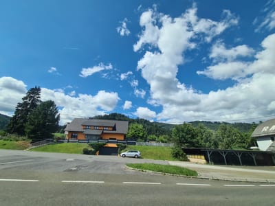The image depicts a street scene in St. Peter ob Judenburg, Austria, under a partly cloudy blue sky. In the foreground, a two-lane asphalt road with white lane markings runs diagonally across the frame. To the right of the road, a paved area with grass borders leads to a covered parking structure with a yellowish wall and a dark roof supported by white pillars. Further back and slightly to the left is a multi-story orange house with a dark gray roof, featuring multiple windows. A silver station wagon is parked on a grassy incline in front of the house, near a dark garage door. A metal staircase with railings ascends the grassy slope to the left of the house. A tall evergreen tree stands to the left of the staircase, and behind the house, a dense forest covers rolling hills that rise into mountains in the distance. A faint white contrail from an aircraft is visible in the upper left quadrant of the sky.