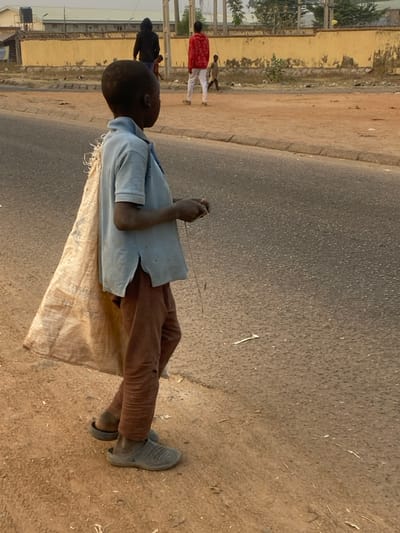 A young boy stands on a street, wearing brown pants, a light blue shirt, and dark grey sneakers. He carries a large tan sack over his shoulder. He appears to be manipulating a thin object in his hands. In the background, blurred figures walk alongside a road. The surrounding environment consists of a mix of asphalt, dirt, and vegetation. Buildings are visible in the distance.