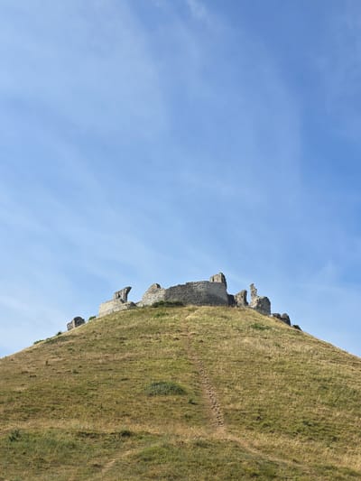 The image depicts the ruins of Corfe Castle, a historic castle located in the village of Corfe Castle, Dorset, England. The castle is situated atop a grassy hill, with crumbling stone walls and structures visible against a clear blue sky with sparse clouds. A well-worn path winds up the side of the hill towards the ruins. The surrounding landscape is characterized by dry, golden grass, suggesting a sunny and possibly dry season. There are no people or animals visible in the scene. The overall impression is one of historical grandeur and natural beauty.