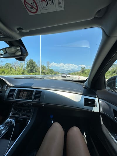 The image is a point-of-view shot from the interior of a car driving on a sunny day. The view is out of the windshield and shows a road with other cars, trees, and mountains in the distance. The dashboard of the car is visible, including the infotainment screen, air vents, and gear shifter. The legs of the driver or passenger are visible in the foreground. The overall scene suggests a journey through a scenic, possibly rural, area of Montenegro, with the specific location of Bratešići potentially being along this route.