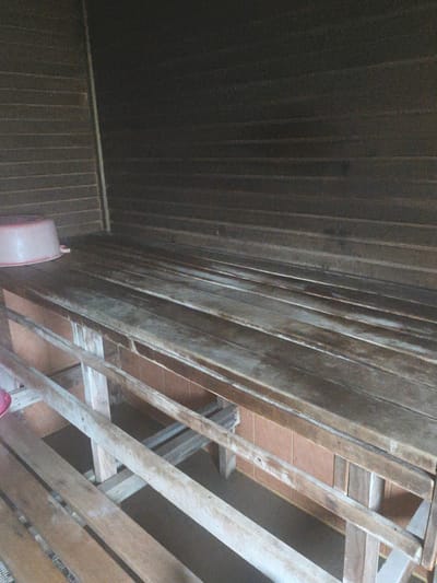The image shows a wooden bench inside what appears to be a sauna or a similar wooden room. The bench is made of several wooden planks. In the background, the walls are also made of horizontal wooden planks. A light pinkish plastic basin is visible on the left side of the bench. The floor beneath the bench is not clearly visible, but there appears to be some tiling or a different material. The lighting is dim, suggesting an indoor environment. The location is identified as Brodi, Latvia.