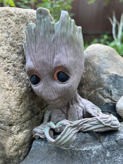A close-up, eye-level shot captures a Baby Groot action figure nestled amongst rocks. The figure, made of grey plastic with green accents on its head and arms, is positioned on a dark grey rock in the foreground. Its large, black eyes are centered, and its twig-like hands are clasped in front of it. To the left, a large, textured tan rock juts into the frame, partially obscuring the figure's head. In the background, out-of-focus greenery and trees suggest an outdoor, natural setting. The lighting is soft, illuminating the scene evenly. There is no discernible indication of a specific location within Angarskie Khutora, Russia; the environment appears to be a garden or park.