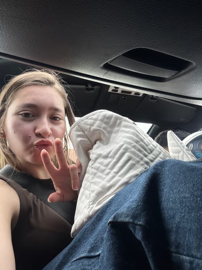 A young person, likely female, with light brown hair, takes a selfie from a low angle inside a car. She is positioned in what appears to be the passenger seat, looking directly at the camera. Her left hand is raised, forming a peace sign with her index and middle fingers, while her lips are playfully pouted. She is wearing a brown-colored top, and a seatbelt strap crosses her shoulder.
To her right, a light-colored, possibly white or cream, quilted garment or bag with a chevron pattern is visible, partially covered by dark blue denim fabric, likely jeans. The background reveals the dark interior of a car, including textured dashboard or door panel surfaces, an air vent, and some control buttons. A cup with partially visible text, including the word "Happy" and "Sleep SMILE", is also faintly visible in the upper right background. The bright light filtering in from outside suggests it is daytime. The overall impression is one of a casual, playful moment captured within a vehicle. While the image is set in Radinovo, Bulgaria, no specific visual cues within the photo itself indicate this location beyond the car interior.