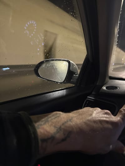 A partial view from inside a vehicle shows the passenger-side door panel, window, and exterior mirror, indicating travel within the Sozina Tunnel in Montenegro. The foreground features a person's right forearm and hand, resting on the dark interior trim. The hand displays visible veins and dark, abstract tattoos on the forearm. Raindrops cover the exterior of the passenger window and the side mirror. Through the wet window, the brightly illuminated, light-colored walls of the tunnel are visible. A reflection of the vehicle's illuminated instrument cluster, specifically a circular gauge with numeric indicators and additional digital readouts, is distinctly projected onto the window glass.