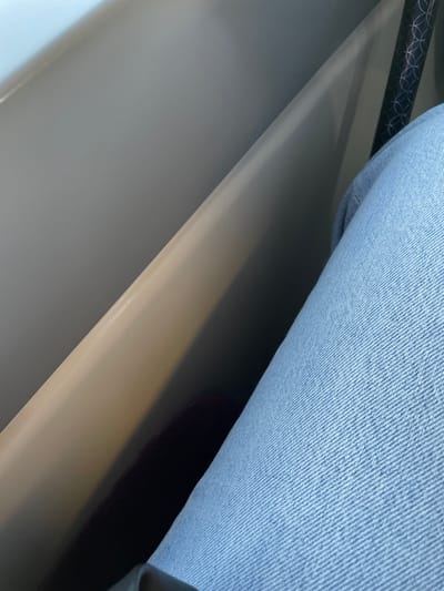 The image captures an interior scene within an unspecified location in Hochfelden, France. The foreground on the right side is occupied by the upper leg and knee of an individual, clad in light blue denim jeans, exhibiting a visible diagonal twill weave. A smooth, light beige or off-white curved partition or panel runs diagonally from the upper left to the lower center of the frame, showing a prominent highlight along its upper edge from a light source originating from the top left. A darker, shaded void is visible behind this partition. In the upper right quadrant, a section of a dark object with a repetitive circular or geometric pattern is present, suggestive of a structural element like a pole or seat support. The scene depicts a static arrangement, implying the individual is seated.