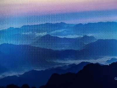 The image displays a gradient of colors from pink and purple at the top to deep blue at the bottom, simulating a mountainous landscape at dusk or dawn. Multiple layers of silhouetted mountains recede into the distance, with the foreground mountains being the darkest. The texture of the image suggests it is a digital display, possibly a computer screen, as individual pixels are discernible. In the bottom right corner of the image, the letter "G" is visible, rendered in white. The scene does not contain any people, actions, or interactions. The location in Reftinsky, Russia, is not evident from the visual information provided in the image.