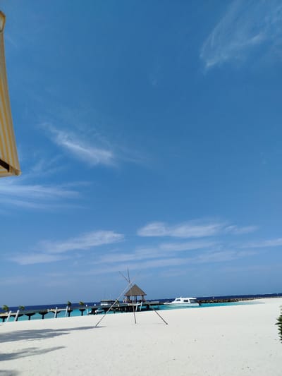 A tranquil scene unfolds on a white sand beach in Kendhoo, Maldives, under a vast, clear blue sky dotted with wispy white clouds. In the foreground, a portion of a yellow and white striped umbrella is visible at the upper left, casting subtle shadows on the sand. Further out, a wooden pier stretches into the turquoise water, its pilings creating a rhythmic pattern. At the end of the pier, a small, thatched-roof hut stands, a beacon of shelter. A sleek white boat is moored near the pier, its presence suggesting leisure and travel. In the middle ground, a unique, X-shaped wooden structure, possibly a decorative wind chime or art installation, stands prominently on the sand. The water transitions from a bright turquoise near the shore to a deeper blue as it meets the horizon. The overall impression is one of serene beauty and relaxation, characteristic of a tropical paradise.