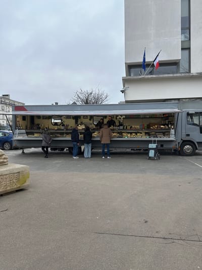 The scene is an outdoor market in Serris, France, set on a paved surface beneath an overcast sky. A grey mobile market truck, with its side extended and open, serves as the central subject. Inside, two vendors operate behind display cases filled with cheeses, baked goods, and bottles. Five customers, dressed in winter coats, face the vendor display; one stands alone on the left, while four others gather closer to the center-right. A blue and black wheeled shopping trolley is parked on the pavement to the right of the truck. In the background, a multi-story, light-grey modern building features prominent windows and two flags: the blue European Union flag and the French tricolor flag. A surveillance camera is affixed to this building's facade. A dark blue passenger vehicle is partially visible behind the market truck on the far left. A light-colored stone block inscribed with 'MBE PAIX' is visible in the lower-left foreground. Bare tree branches are visible in the mid-ground.