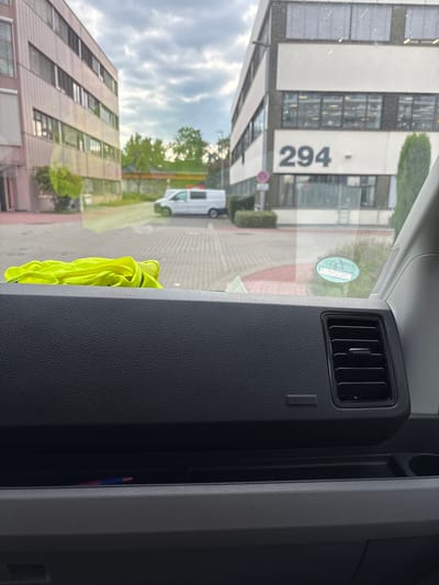 The image, taken from inside a vehicle, shows a view through the windshield of two modern office buildings. The building on the left is lighter in color with rows of windows and blinds. The building on the right is darker, with a prominent sign displaying the number "294". A white van is parked in the lot between the buildings. The foreground of the image reveals a black textured dashboard with an air vent and an "AIRBAG" label, as well as a bright yellow folded piece of fabric. The environment appears to be an office park or industrial area in Mannheim, Germany, under an overcast sky.