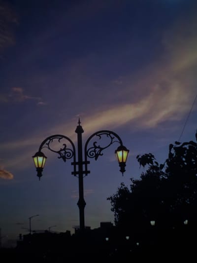 A silhouette of a streetlamp with two illuminated bulbs stands against a twilight sky with wispy clouds. The streetlamp is ornate, with curved metal arms and decorative scrollwork. In the background, silhouetted trees and buildings are visible, along with several other distant, dimly lit streetlights. The scene appears to be an outdoor urban setting, possibly in the city of Masis, Armenia. The overall atmosphere is calm and serene, characteristic of dusk.