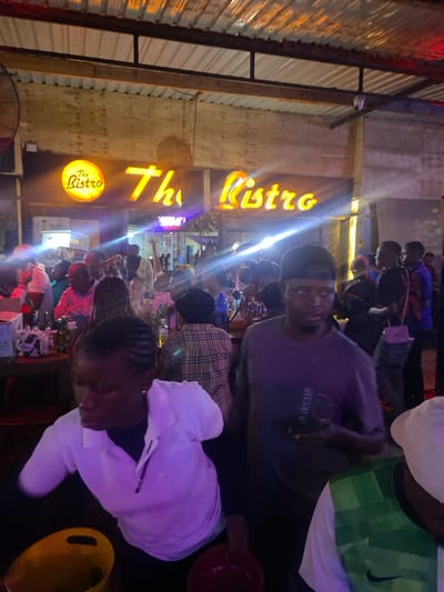 The image shows the interior of a bar named "The Bistro" located in Dodoma, Tanzania (latitude: -6.157, longitude: 35.724). The bar is brightly lit with neon signs. There are several people inside the bar, standing and sitting, some interacting with each other. Some people are at the bar counter, which is covered with a wooden top. The counter is illuminated with bright lights. The bar has a wooden ceiling with exposed beams. There are a few yellow buckets on the floor. A person wearing a white shirt with a light purple hue is bending over, presumably cleaning something. A man in a grey shirt is standing in the center of the image, holding a black phone in his left hand. His right hand is at his side. He is looking at his phone. A man in a plaid shirt is to his left, and another man in a green and white shirt is to his right. The man in green and white has a white cap on his head and is looking down. A group of people are in the background, some of whom are chatting and some of whom are holding drinks. The people are all African, dressed in casual clothes. The scene appears to be taken at night, and there is a lot of light coming from the neon signs. The image was taken at a slight angle from a lower level.