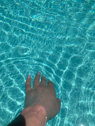 A person's hand is submerged in the clear, turquoise water of a swimming pool, with visible tile lines on the pool floor. The sunlight creates dappled patterns of light and shadow on the water's surface, giving it a shimmering effect. The hand appears to be reaching out or touching the water, with manicured fingernails visible. The scene is set in a swimming pool, possibly in Mardakan, Azerbaijan, suggesting a warm, outdoor environment.