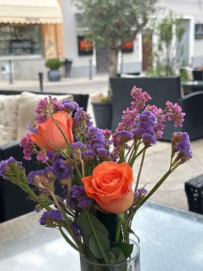 A close-up shot focuses on a bouquet of orange roses and purple statice flowers in a clear glass vase. The flowers are arranged in the foreground, with their stems reaching upwards. In the background, an outdoor cafe setting is visible. Chairs and tables of the cafe are blurred, with an olive tree also out of focus. A building with an awning and a sign that reads "BAZA" is in the distance. The overall scene suggests a relaxed outdoor dining or cafe experience in a Mediterranean town, likely Carbonaxa or Villasimius, Italy, given the architectural style and vegetation.