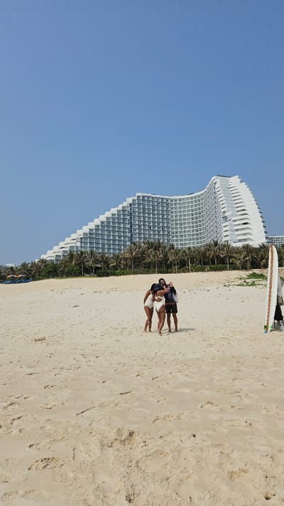 A couple stands on a sandy beach in Bac Cam Ranh, Vietnam. The woman is wearing a white two-piece swimsuit, while the man is wearing a dark shirt and dark shorts. He appears to be taking a photograph. A large white building is in the background, next to a row of palm trees. There is a surfboard at the top of the frame. The sky is clear blue.