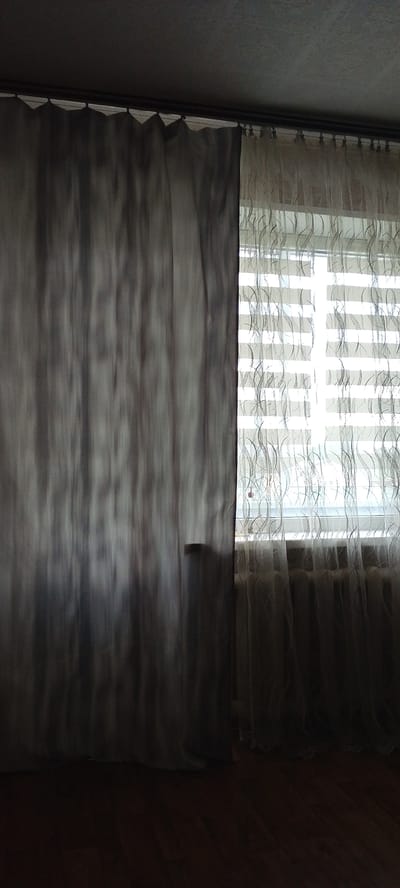 The image shows a window with curtains in Lyudinovo, Russia. The left side of the window is covered by a dark gray curtain, and the right side is covered by a sheer white curtain with a wavy pattern. The window itself appears to be a standard-sized residential window, and through the sheer curtain, blinds can be seen. The floor beneath the window is made of dark wood planks. The ceiling above the curtains is textured and appears to be painted a dark gray. The overall lighting suggests it is daytime, but the room is dimly lit, possibly due to heavy curtains or overcast weather. There is a small, light-colored rectangular object protruding slightly from the dark gray curtain, located roughly in the middle of the curtain's height. No people or animals are visible in the image.