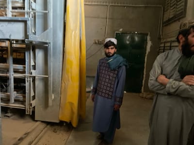 The image depicts an indoor scene within a facility in Shuyene Vosta, Afghanistan. Two adult males are present. On the left, a man with a dark beard and mustache stands facing slightly right of the camera. He wears a white skullcap, a blue scarf draped over his left shoulder, and a patterned dark vest over a light blue tunic. To his right, partially visible, is another man with a dark beard, wearing a light grey tunic with arms crossed.
The environment features a large, multi-tiered grey metal structure on the left, possibly an industrial racking system or machinery, extending from the foreground to the background. Adjacent to this structure on the right is a tall, vertical yellow fabric barrier or curtain. In the background, behind the first man, a dark green double door is visible within an unpainted or unfinished concrete wall. Electrical wiring is affixed to the wall above the door. The concrete floor appears dusty and exhibits tracks or grooves. No specific actions or interactions are observed beyond the individuals standing.