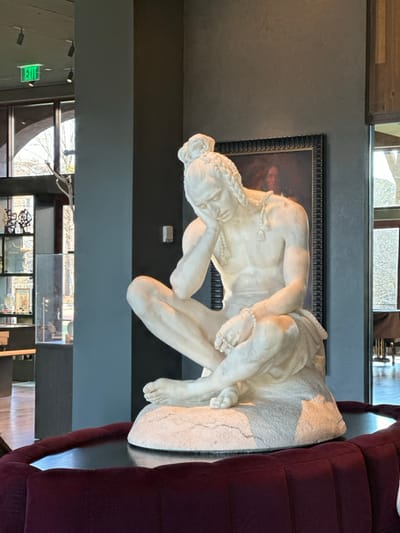 A marble sculpture of a reclining nude figure rests atop a gray surface next to a burgundy couch. The figure is facing left, with head resting on a hand. Behind the sculpture is a framed picture on a gray wall. Windows are visible at the bottom and top of the frame, reflecting sunlight. A green "EXIT" sign is visible through one of the windows.