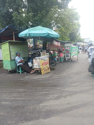 The image shows a street scene in Purwokerto, Indonesia, located at the coordinates -7.417 latitude and 109.249 longitude. A man sits on a chair in front of a food stall, waiting for customers. The stall is under a large blue umbrella and is selling "Cikur Timun Nyida", which appears to be a type of food. In the background, there are other stalls and people walking by. One of the stalls is selling "Es Pisang Hijau", which may be a type of banana-based dessert. The environment is urban, with the scene taking place on a paved road. The sky is overcast with clouds. Several people are walking down the street. There are various food stalls set up along the street with people standing around. The man sitting is likely a vendor. The image is taken from an angle that shows the street, stalls, and people walking by. The road is wet from rain.
