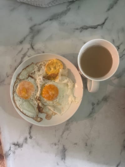 The image depicts a breakfast scene on a marble-patterned surface. A white plate holds three fried eggs and a cracker. To the right of the plate is a white mug filled with a light brown beverage, likely coffee. The background shows a white textured fabric. The scene is indoors, with artificial lighting. The specific location in the city of Şu, Kazakhstan, cannot be determined from the visual information provided in the image.