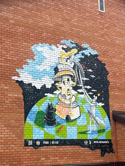 This media file is a photograph of a mural painted on a brick wall in Mezhdurechenskiy, Russia. The mural is a colorful and abstract depiction of space and Earth. The left side of the mural shows a swirling galaxy with stars and nebulae, rendered in black, white, and blue. Emerging from this cosmic scene is a stylized, metallic object that resembles a complex piece of machinery or a spacecraft, with gears and pipes visible. Yellow and orange hues highlight parts of this object.
The right side of the mural transitions to a checkered pattern of green and blue, representing the Earth or a similar planet. Across this celestial landscape, a white, flowing form resembling a ribbon or a wing stretches from left to right. A silhouette of a bird or an angel with outstretched wings is subtly integrated into this form. There is also a small, orange carrot depicted on the green checkered section. A black object that looks like a vintage camera or projector is positioned in the lower right quadrant of the Earth section.
The mural is painted on a red brick wall. In the upper left corner of the photograph, a white ventilation grate is visible on the brick wall. On the far right edge of the mural, there is a vertical black strip containing white text and symbols. The text reads "ИГРА НАЧАЛАСЬ" which translates to "The Game Has Begun" in Russian. Below this are several small icons and the hashtag "#USARTCO". A QR code is also present at the bottom of this black strip.
The overall impression of the mural is one of transition and exploration, with elements of the abstract and the tangible. The lighting suggests it is daytime, with diffused light, but no specific time of day or weather conditions are evident. The mural appears to be located outdoors, on the side of a building.
