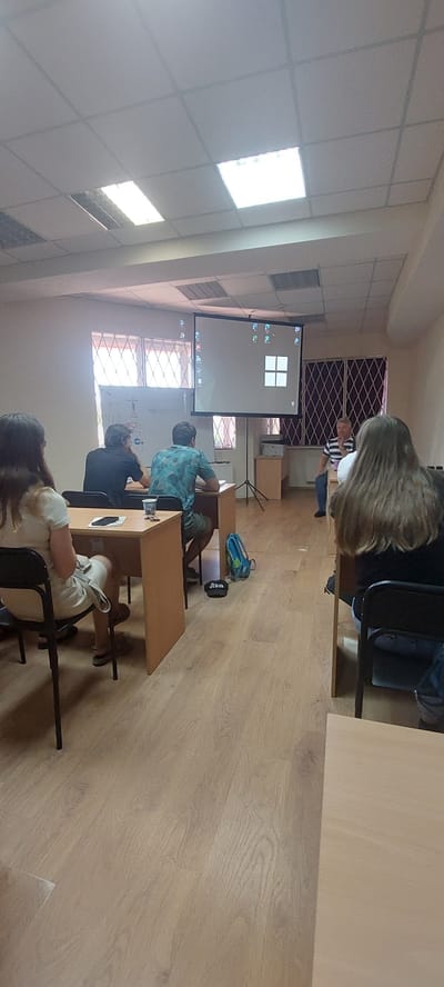 The image shows a classroom or lecture hall setting in Tetiiv, Ukraine. A projector screen displays a Windows operating system interface with four square windows arranged in a grid. Several individuals are seated at desks facing the screen. On the left, a person with long brown hair is seated at a desk. In the middle, two individuals are seated at desks, one wearing a black shirt and the other a patterned blue and green shirt. A blue backpack and a black cap with white lettering are on the floor near the middle desks. To the right, a person in a white and blue striped shirt is standing and speaking, gesturing with their hands. Another person with long brown hair is seated at a desk on the far right, facing away from the camera. The room has fluorescent lighting on the ceiling and wooden floors. Windows with barred coverings are visible on the back wall.