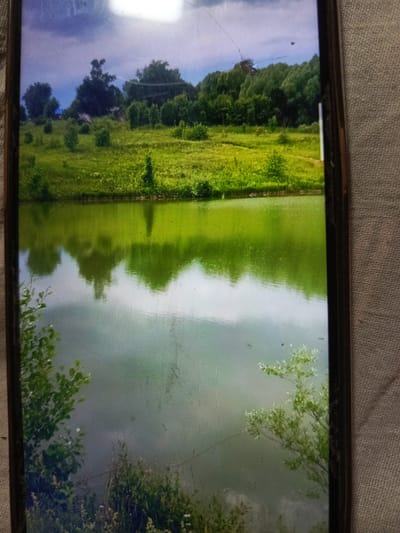 The image displays a serene landscape scene, likely a rural setting, featuring a body of water with reflections and a grassy hillside dotted with trees and a few small structures. The sky above is partly cloudy, with sunlight casting a bright glow. The overall impression is one of natural beauty and tranquility. However, there is no information within the image itself or the provided context to link this scene to the city of Svoboda, Russia, or any specific urban location. The image is a photograph of a natural environment.