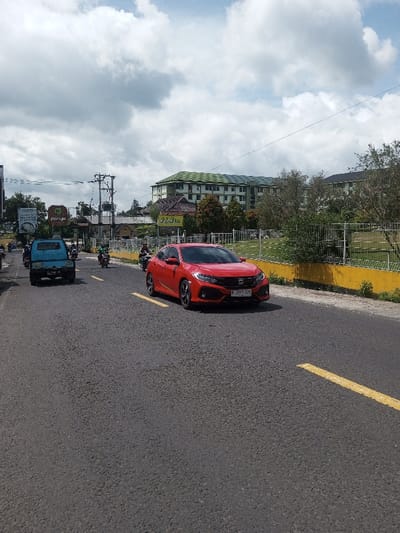The scene depicts a daytime street view featuring vehicular traffic under an overcast sky. A red Honda Civic sedan, displaying Indonesian license plate "B 201 F 57G," occupies the right side of the asphalt road. To its left, a light blue pickup truck, an older model, moves in the same general direction. Several motorcycles, some with riders, are also visible on the road. Yellow dashed lines mark the lanes. On the right, a yellow concrete barrier separates the road from a grassy area secured by a metal fence. In the background, a large multi-story building with a light green roof and light-colored walls stands prominently. Further left in the background, power poles, smaller buildings, and commercial signage, including a green sign bearing the text "ponyo," are present. The license plate "B 201 F 57G" identifies the vehicle as registered in Jakarta, Indonesia, which contradicts the provided location of Gubbisamamidi, India.