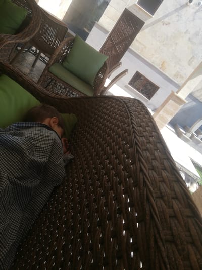A young boy wearing a checkered shirt sleeps on a green cushion on a wicker chair. The chair is on a patio with other wicker furniture. The ceiling above appears to be a pergola with a translucent roof. The scene is brightly lit by sunlight, casting shadows on the floor. The boy is lying on his side, facing away from the camera, with his head resting on the cushion. His eyes are closed, and his breathing is likely slow and steady as he sleeps. The wicker chair is intricately woven, with a thick braid forming the armrest and back. The green cushion is plump and firm, providing a comfortable surface for the boy's rest. The patio appears to be an outdoor dining or lounging area, possibly in a restaurant or cafe. The overall impression is one of peace and tranquility, with the boy enjoying a nap in a comfortable setting.