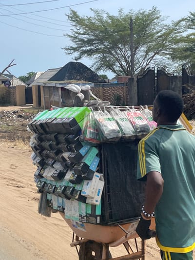 In Ikoga, Nigeria, a man in a green and yellow shirt is pulling a cart. The cart is heavily loaded with various boxed and packaged items. A building is visible in the background. The scene is outdoors, and the ground appears to be sandy or dirt-covered.