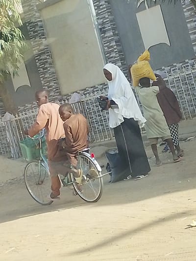 The image depicts a daytime outdoor scene featuring several individuals on an unpaved, dusty path or street, against a backdrop of a building and some foliage.
In the left foreground, two young males are interacting with a bicycle. One, wearing a light brown or orange long-sleeved top and pants, is standing next to the bicycle, holding its handlebars. The bicycle is light blue or green with a green plastic basket mounted on the front. A second young male, dressed in a similar light brown top and darker pants, is seated on the back of the bicycle, facing towards the left, with light-colored sandals on his feet. The bicycle has a red reflector on its rear fender.
To the right of the bicycle, a group of three females is walking. The most prominent among them is a woman or older girl wearing a white hijab that covers her head and shoulders, and a long dark skirt. She is holding a dark, possibly rectangular object or small bag in her hands. Next to her, a younger individual, possibly a child, is seen from behind, wearing a light green long garment and expertly carrying a large, bulging yellow sack or bag on their head. Further to the right and slightly behind, another person, also appearing to be young, is walking, wearing a dark top and patterned (possibly checkered) pants or skirt. All the walking individuals appear to be wearing sandals or flip-flops.
The background features a building with a textured wall, possibly made of grey and black tiles. A white metal fence or gate runs in front of this wall. On the far left, some green leaves from a tree or bush are visible, indicating vegetation in the area. The ground is dry, unpaved, and light brown, suggesting a sandy or dusty environment. The bright, natural lighting and clear shadows indicate it is daytime, likely mid-morning or afternoon. No visible text is present in the image.