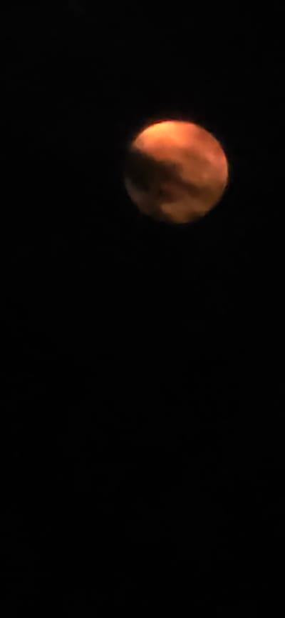 A full moon, appearing orange due to atmospheric conditions, is visible in a dark night sky. Clouds partially obscure the moon. The image is a vertical shot. There is no discernible city or specific location identifiable within the frame.