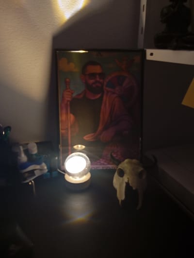 A small glass orb lamp illuminates a dark room, casting a warm glow on a nearby table. On the table, there is a framed picture of a man with a beard, sunglasses, and a long staff. He is wearing a dark shirt and a cloak draped over his shoulders. The picture is set against a background of blue and purple tones. The lamp is casting a reflection of the picture. To the right of the lamp, a dried animal skull with horns sits on the table. The table appears to be made of wood and is set against a white wall. The room is sparsely decorated, and there are no other people present. The room is located in the city of Dunajská Lužná, Slovakia, at latitude 48.074 and longitude 17.273. A shelf with white shelves on the right side of the picture, behind the table, is out of focus. There are objects on the shelves, including a dark object, possibly a sculpture, and a piece of yellow paper, but the objects are obscured by the blur.