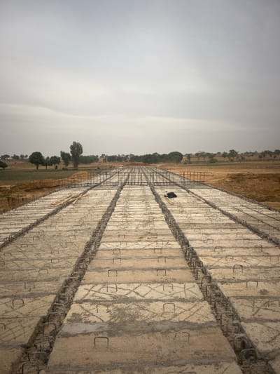 A landscape features a long, concrete structure extending into the distance. The structure is divided into segments with steel reinforcing bars visible. A lone figure stands atop it. The sky is overcast, creating a muted color palette. Adjacent to the structure is a field with scattered trees, adding a rural element to the scene.